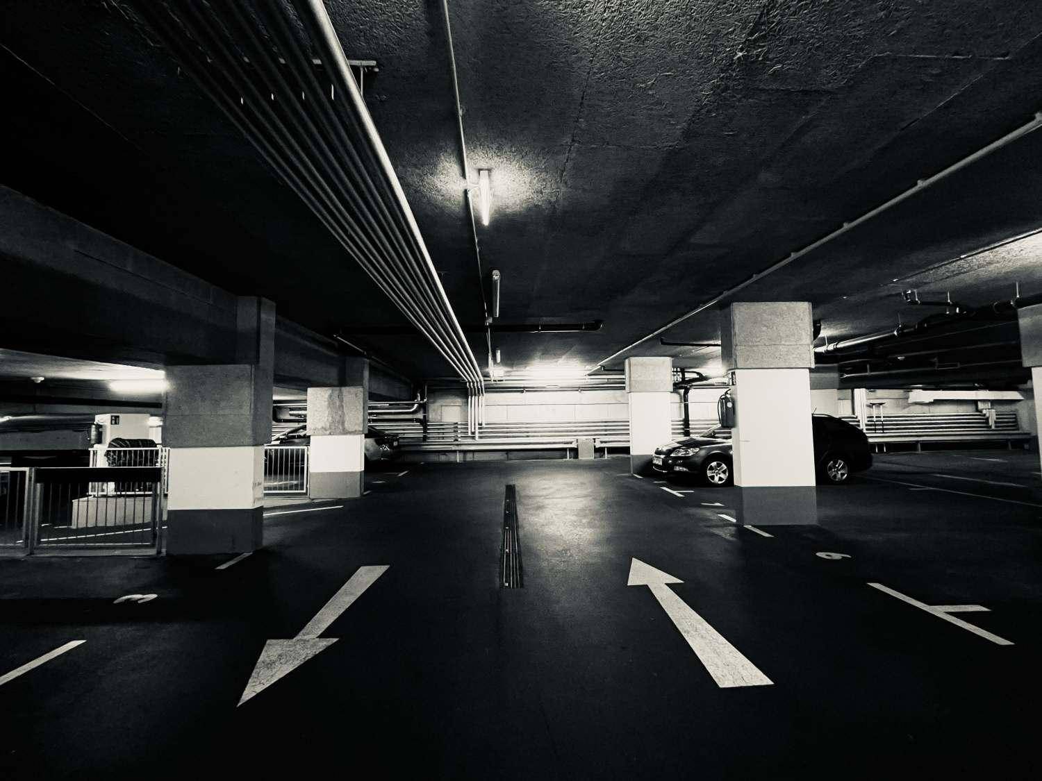 Schwarz-Weiß-Aufnahme einer Tiefgarage mit Betonstrukturen, Rohren und Richtungspfeilen auf dem Boden.