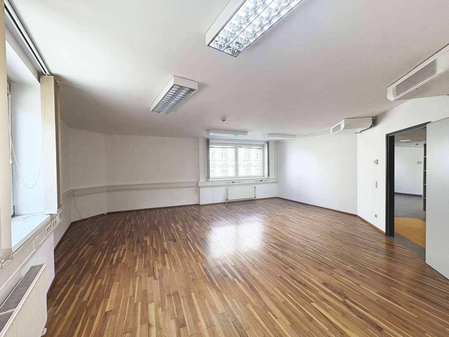 Helles Büro mit Holzparkettboden, mehreren Fenstern und integrierten Lüftungseinheiten an der Decke.