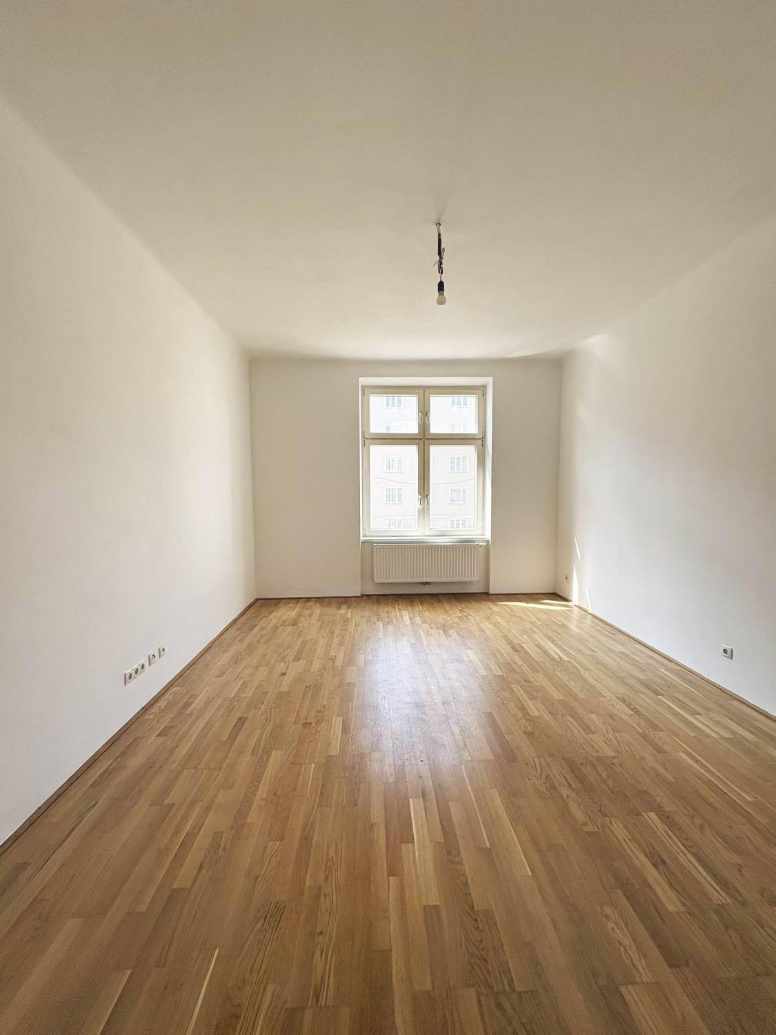 Leerer Raum mit glänzendem Parkettboden und einem großen Fenster, das den Raum mit Licht füllt.