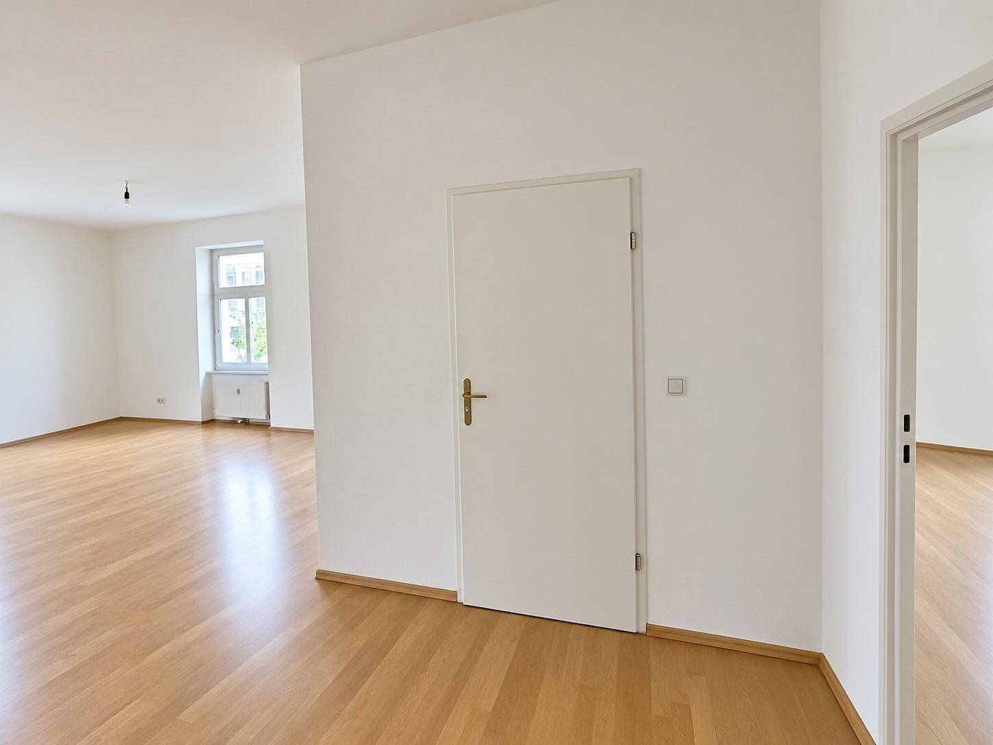 Heller Flur mit weißer Tür und Blick in ein geräumiges Zimmer mit Parkettboden.
