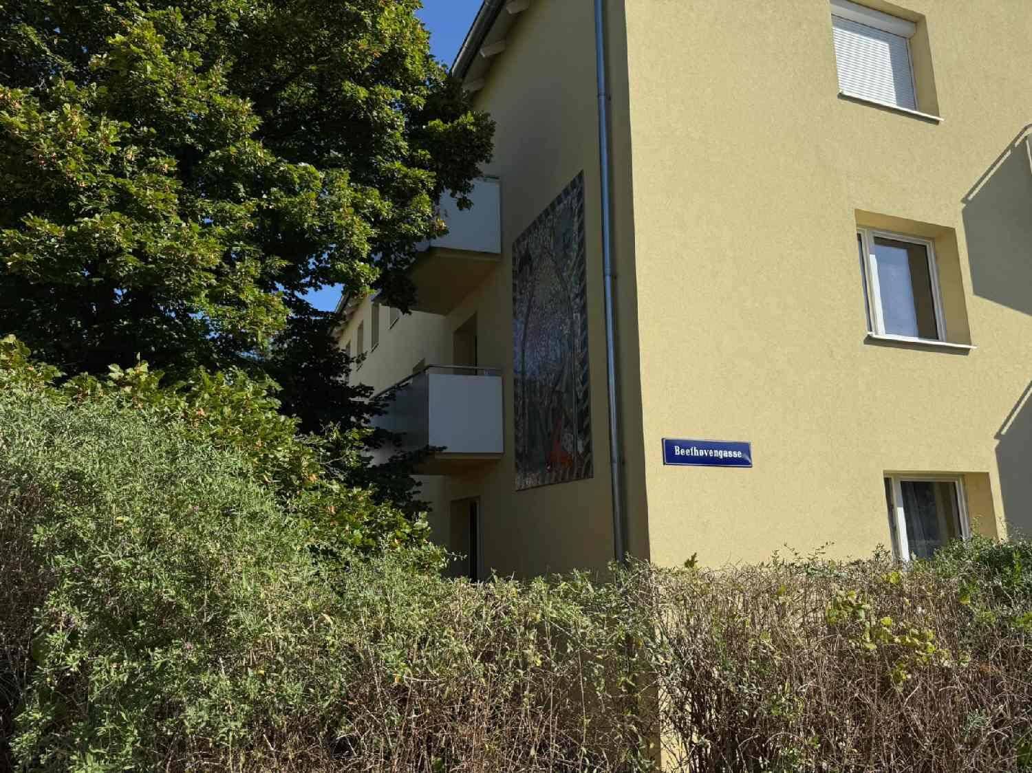 Seitenansicht eines Gebäudes mit gelber Fassade, Balkonen und Straßenschild.