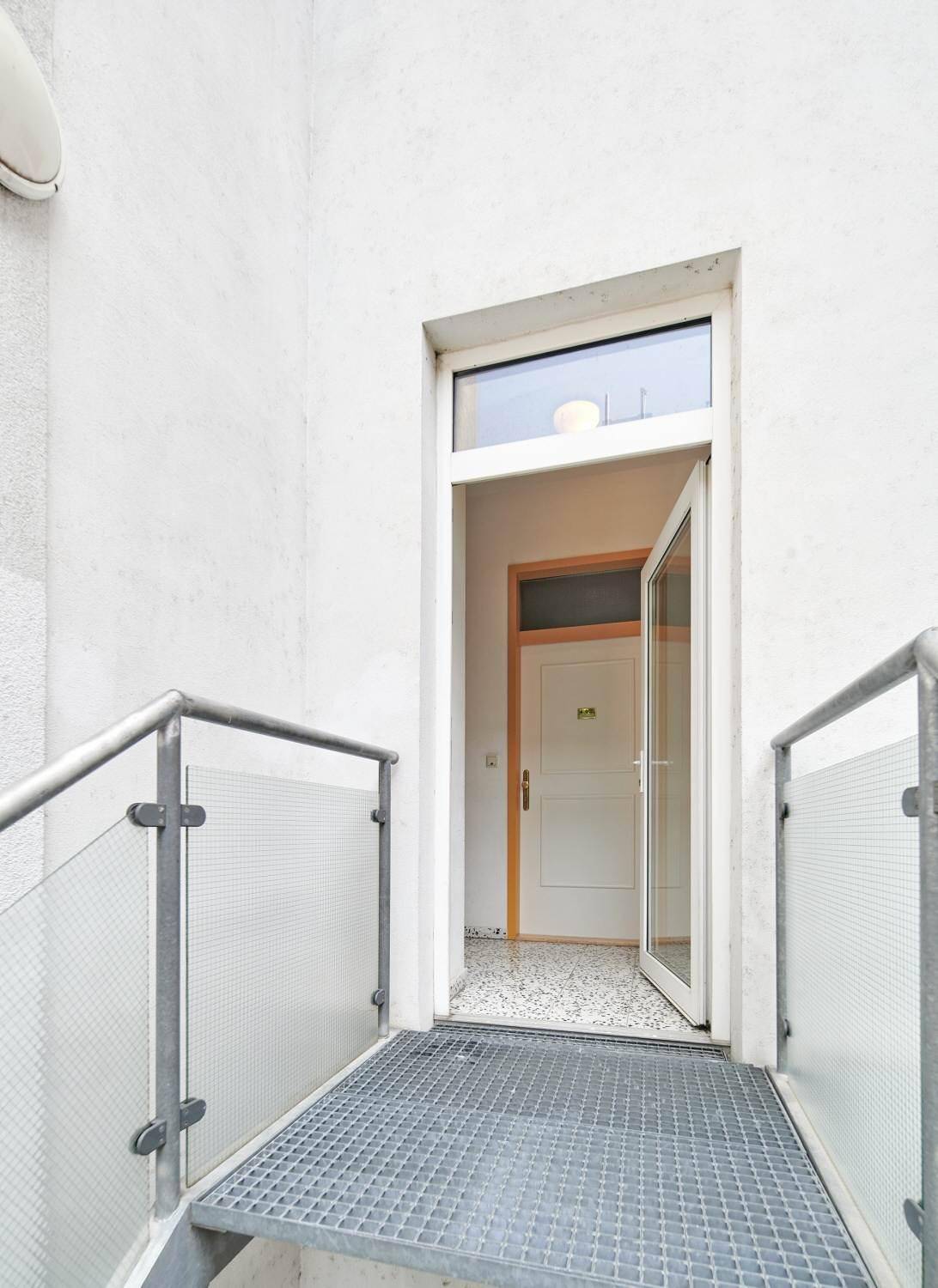 Eingangsbereich mit Metallrampe und Glasgeländer, der zu einer Wohnungstür führt.