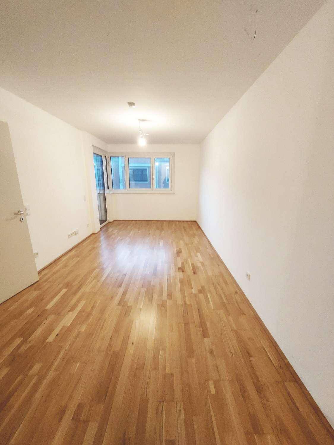 Heller, leerer Wohnraum mit Parkettboden und Fensterfront, die viel Tageslicht hereinlässt.