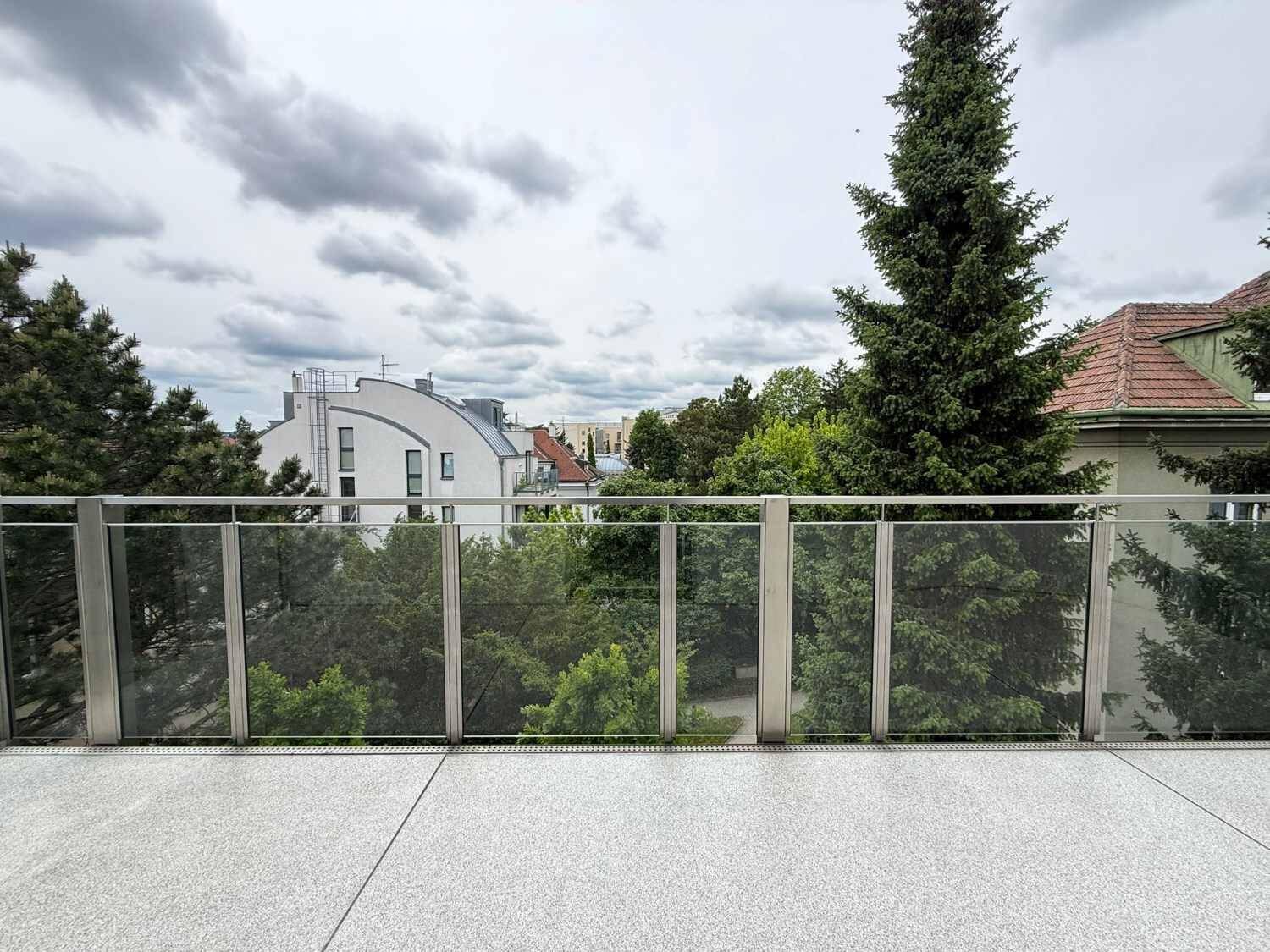 Breiter Balkon mit Glasgeländer und Ausblick auf grüne Bäume und die Umgebung.