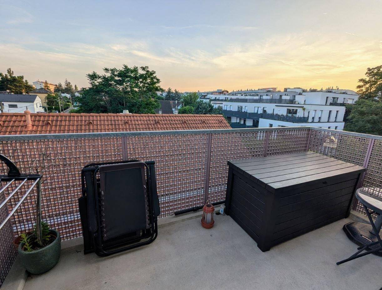 Balkon mit Stauraumbox und Blick über die Dächer der umliegenden Gebäude bei Sonnenuntergang.