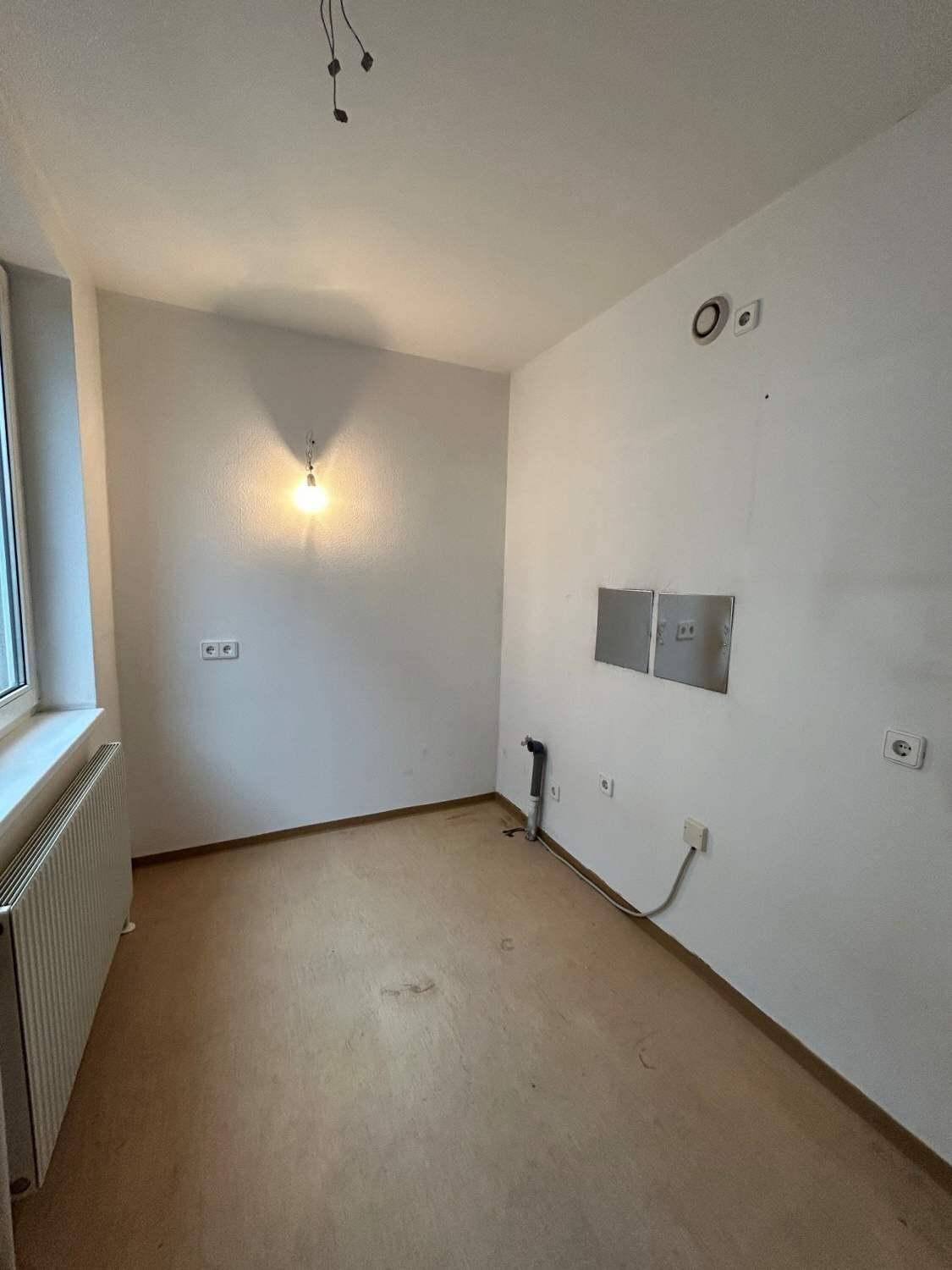 Leerer Raum mit Anschlüssen für eine Küche und einem Fenster, bereit für die Installation einer Einbauküche.