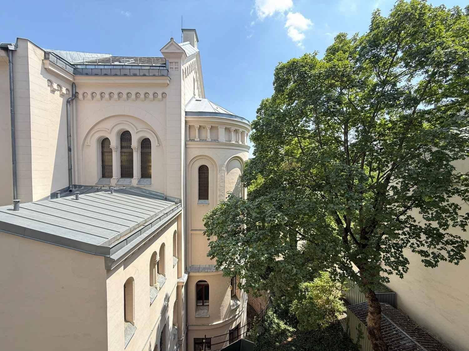 Historisches Gebäude mit kunstvollen Details und einem großen, grünen Baum im Vordergrund unter blauem Himmel.