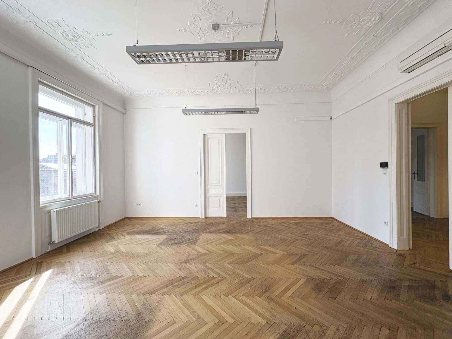 Großes, helles Zimmer mit klassischem Fischgrätparkett und Stuckdecke, ausgestattet mit modernen Leuchten.