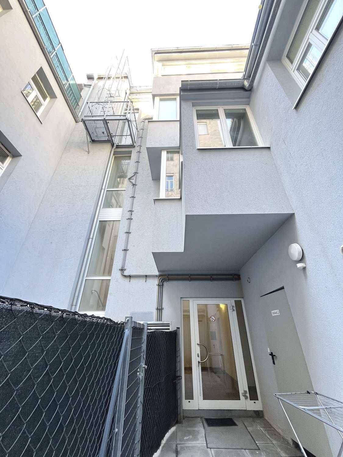 Rückansicht des Gebäudes mit Fenstern, einem externen Treppenaufgang und einem Zaun im Vordergrund.