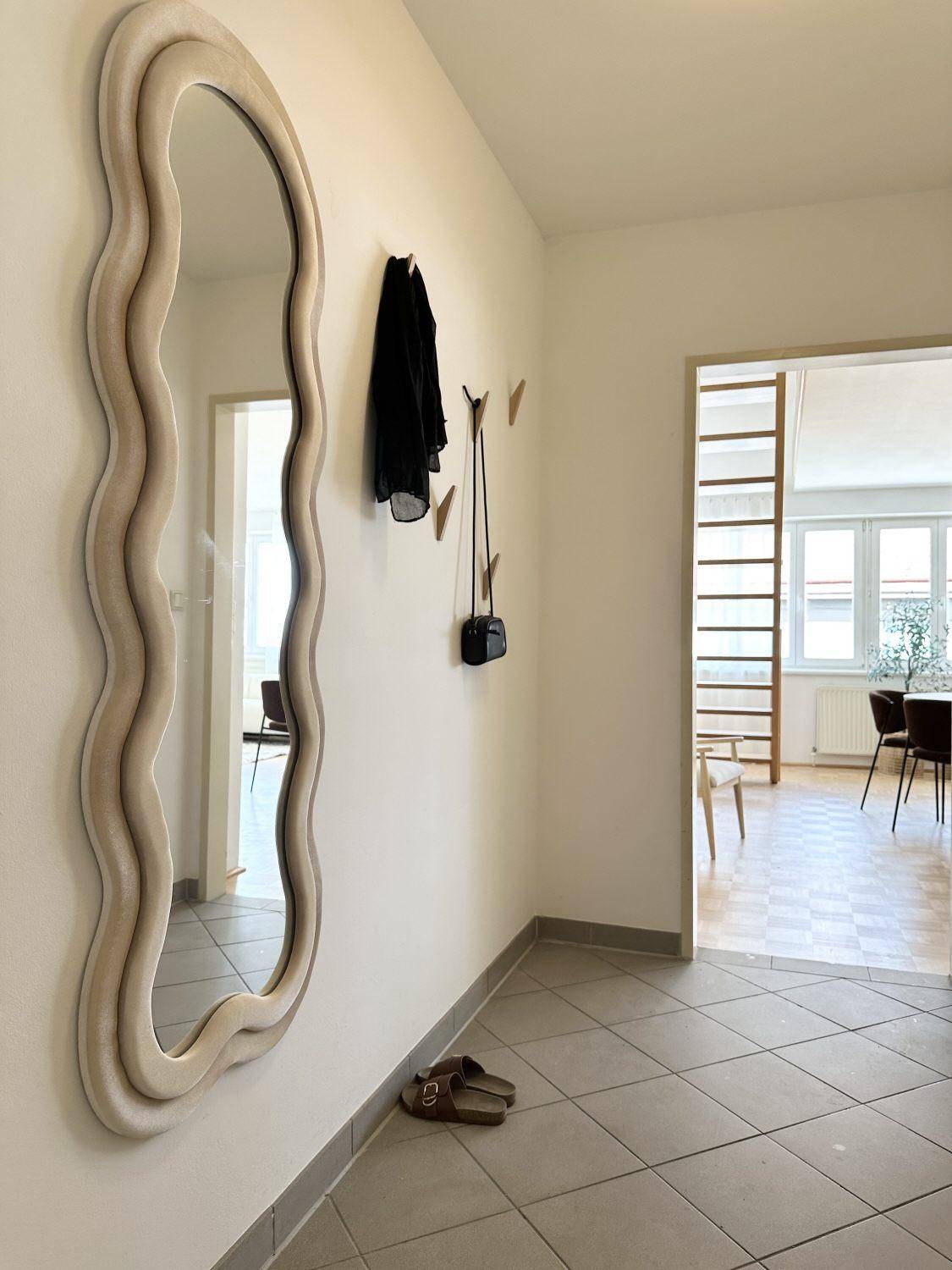 Heller Flur mit modernem Wellenspiegel, Garderobe und Fliesenboden, der zu anderen Räumen führt.
