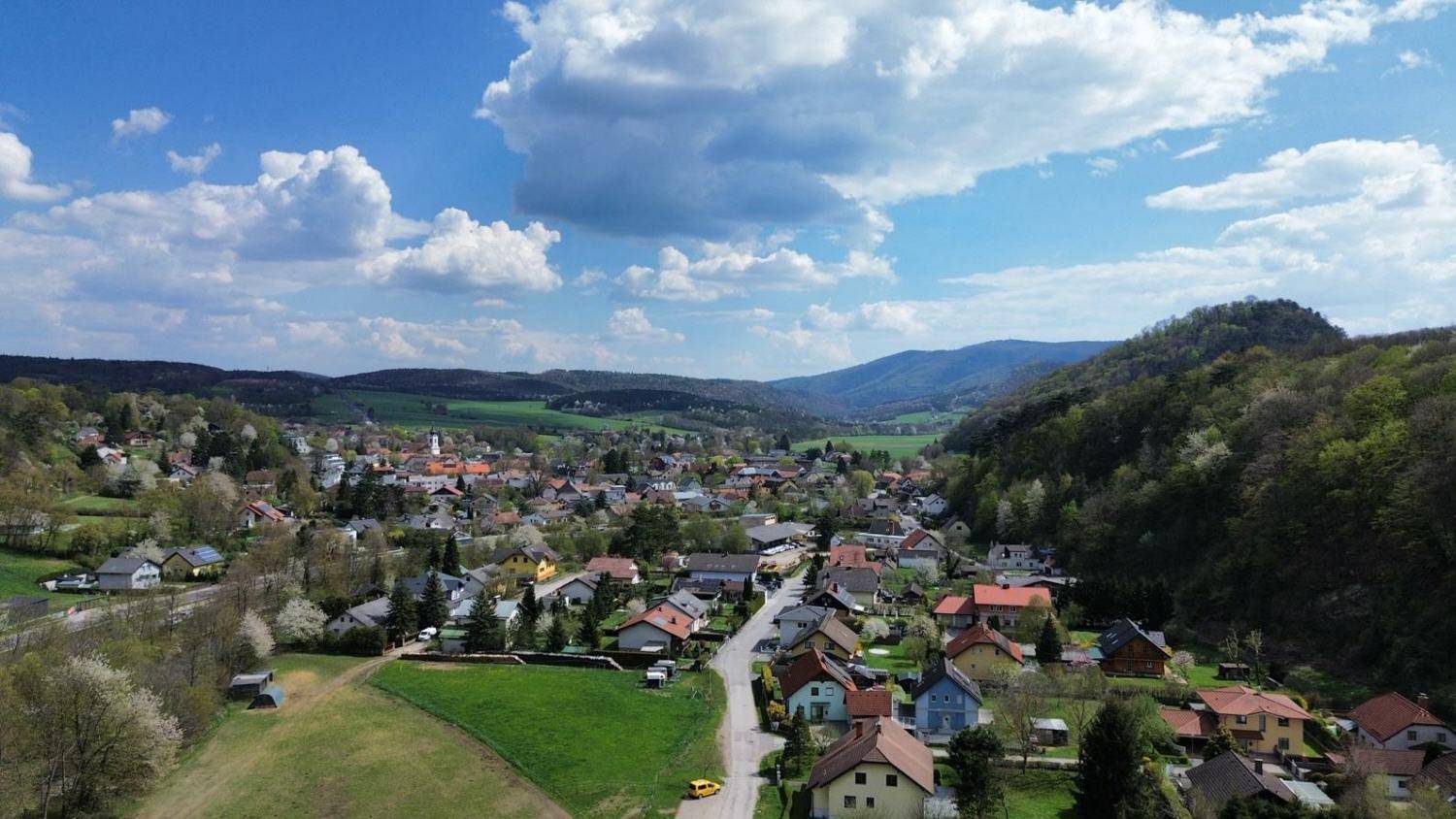 Weitläufige Luftaufnahme eines malerischen Dorfes in einem Tal, umgeben von grünen Hügeln und Wäldern.