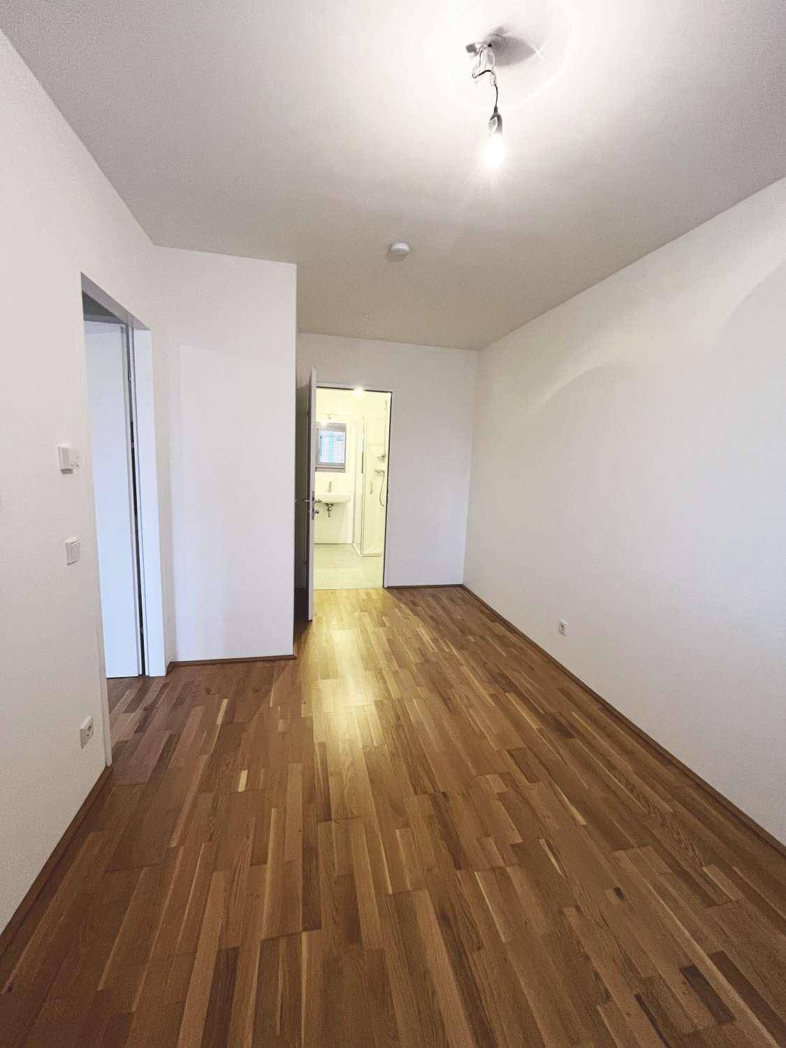 Heller Raum mit Parkettboden und weißen Wänden, mit Zugang zu einem Badezimmer.