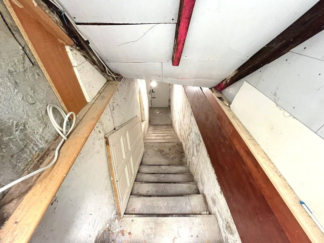 Enge Kellertreppe mit rohen Wänden und provisorischer Beleuchtung, die nach unten führt.