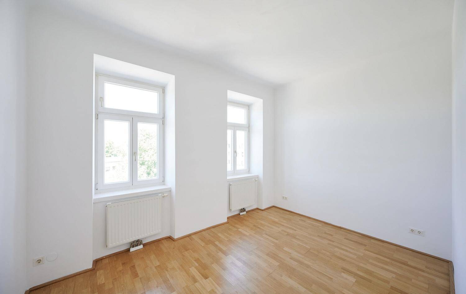 Leeres Schlafzimmer mit hellem Parkettboden und zwei Fenstern, bereit zur Einrichtung.