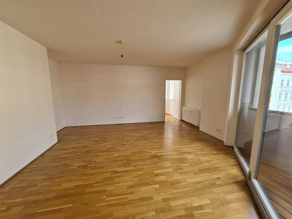 Geräumiges Wohnzimmer mit Parkettboden und großen Fenstern, die Zugang zum Balkon bieten.