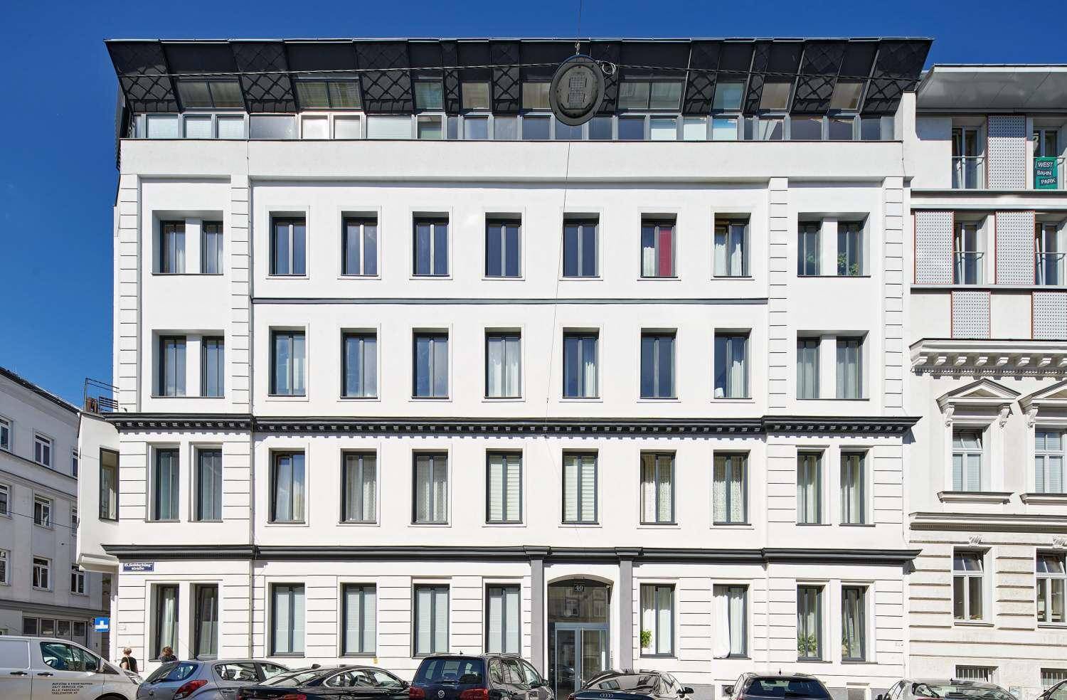 Frontansicht eines modernen Gebäudes mit weißer Fassade, vielen Fenstern und Dachaufbau.