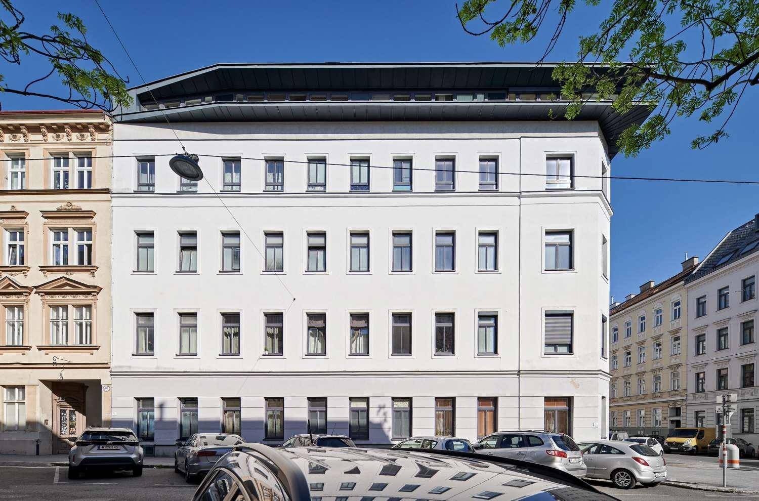 Ansicht der modernen Gebäudefassade mit zahlreichen Fenstern und geparkten Fahrzeugen auf der Straße.