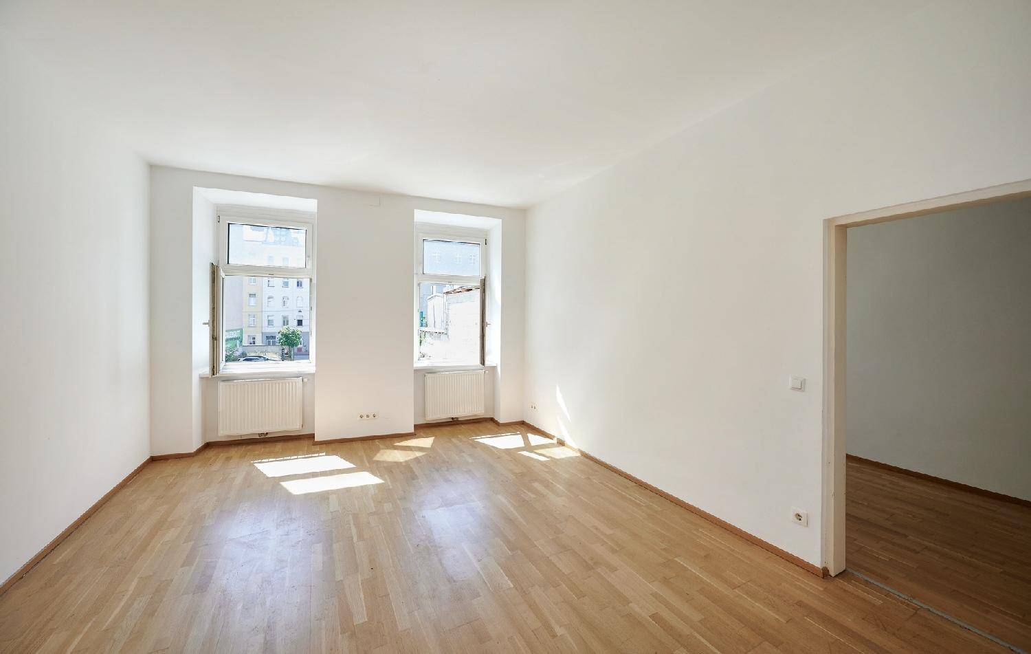 Geräumiger, heller Raum mit Parkettboden und zwei Fenstern, die viel Tageslicht hereinlassen.