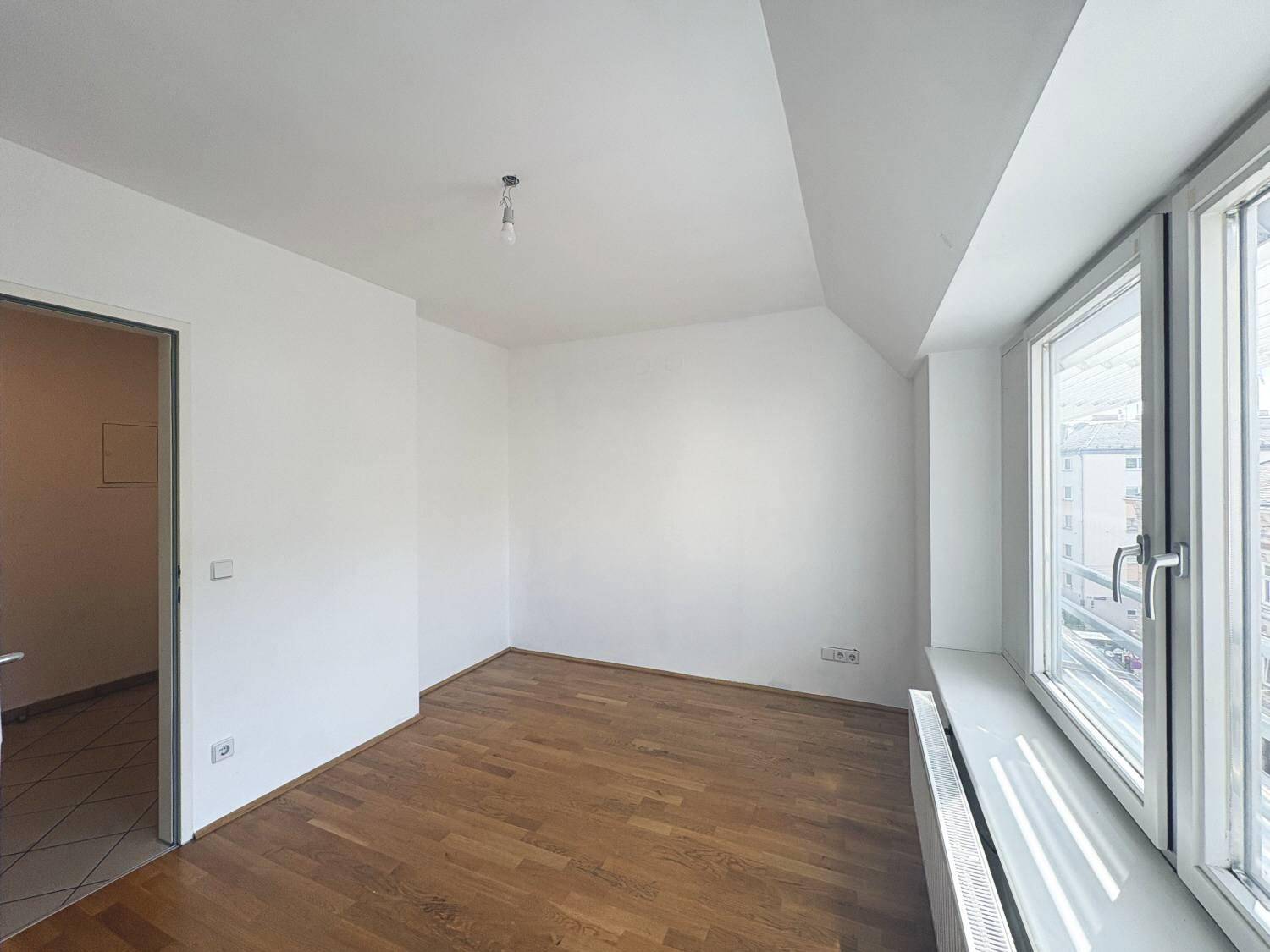 Helles Zimmer mit Holzboden und großem Fenster, bietet Zugang zu einem weiteren Raum.