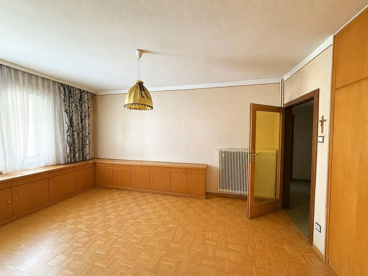 Heller Raum mit Parkettboden, Einbauschränken und einem Fenster mit Heizkörper darunter.
