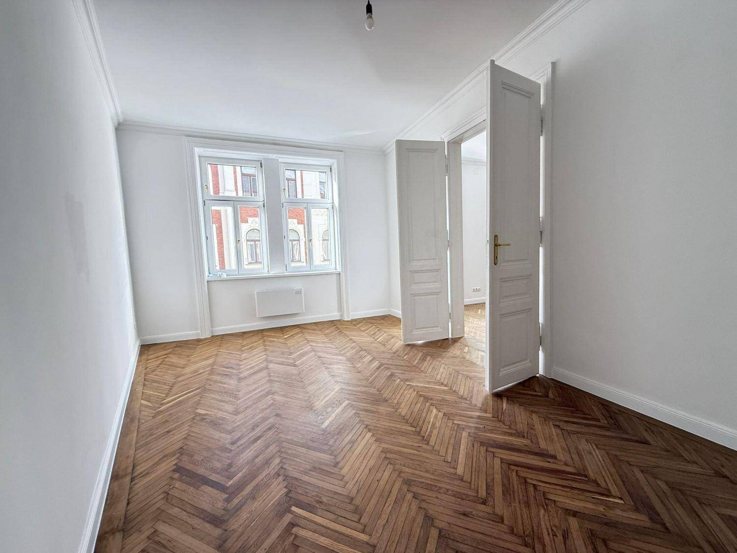 Geräumiges Zimmer mit klassischem Fischgrätparkett und hohen Doppeltüren, die in den nächsten Raum führen.