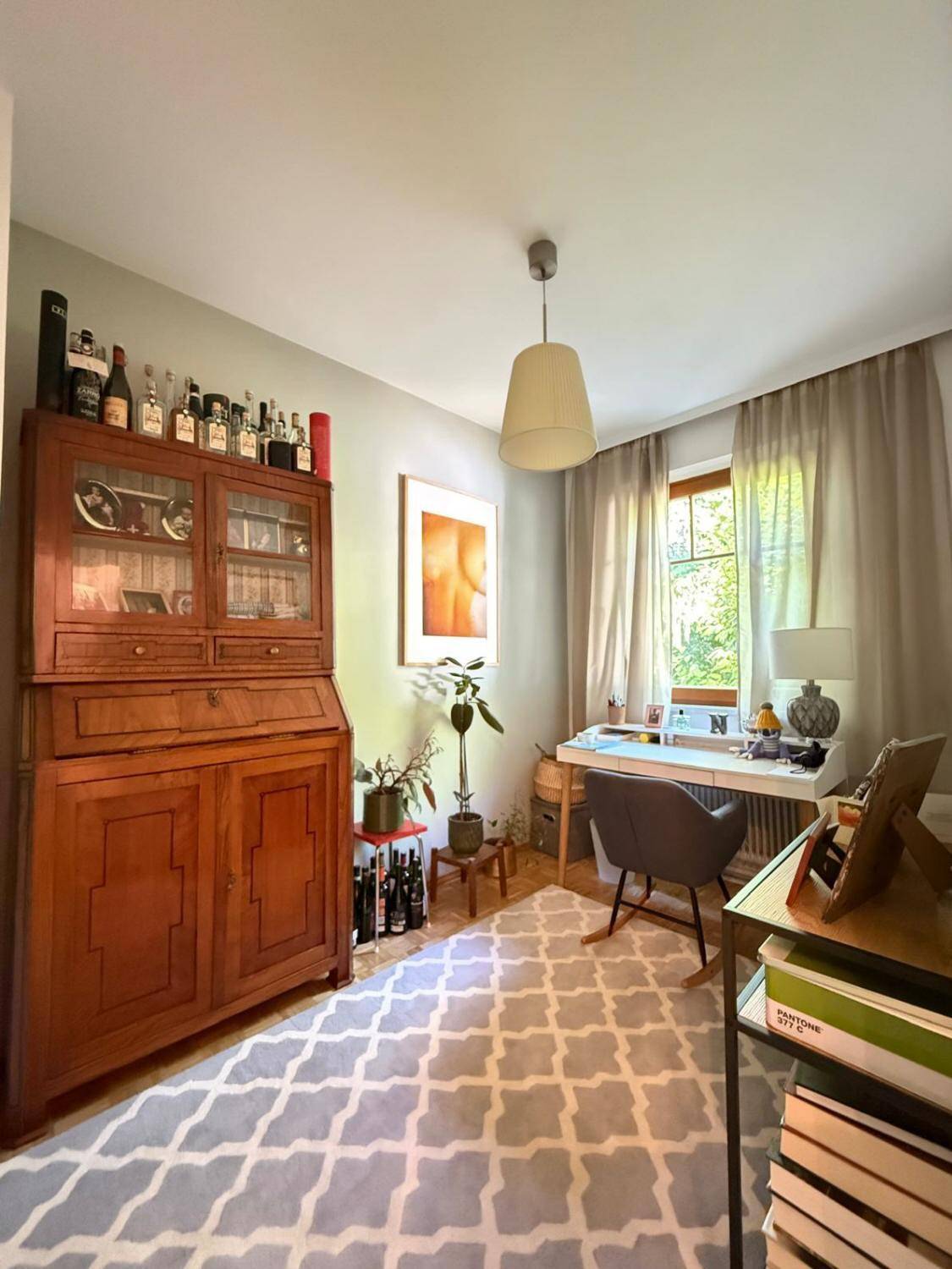 Arbeitszimmer mit Holzschrank, Schreibtisch am Fenster und gemustertem Teppich.