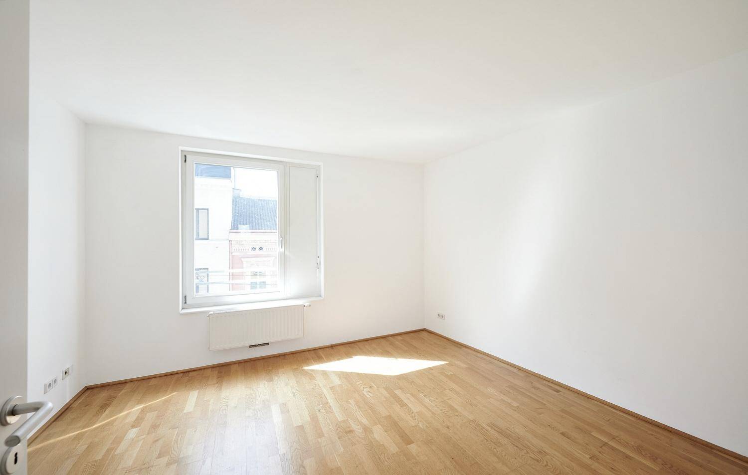 Helles Zimmer mit Parkettboden und einem großen Fenster, das viel Licht hereinlässt.