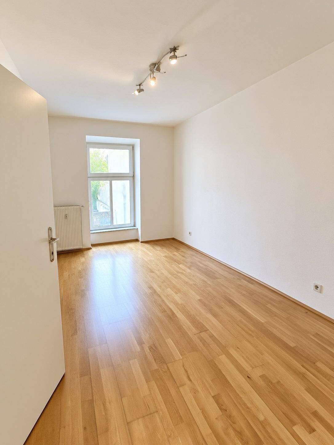 Helles Zimmer mit Holzboden und einem Fenster, das den Raum mit Sonnenlicht durchflutet.