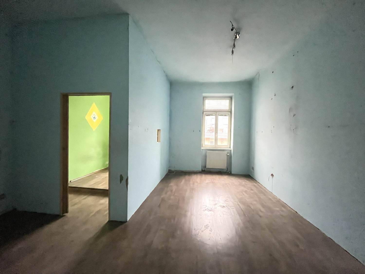 Leerer Raum mit hellblauen Wänden, Holzboden und einem Fenster, das Tageslicht hereinlässt.