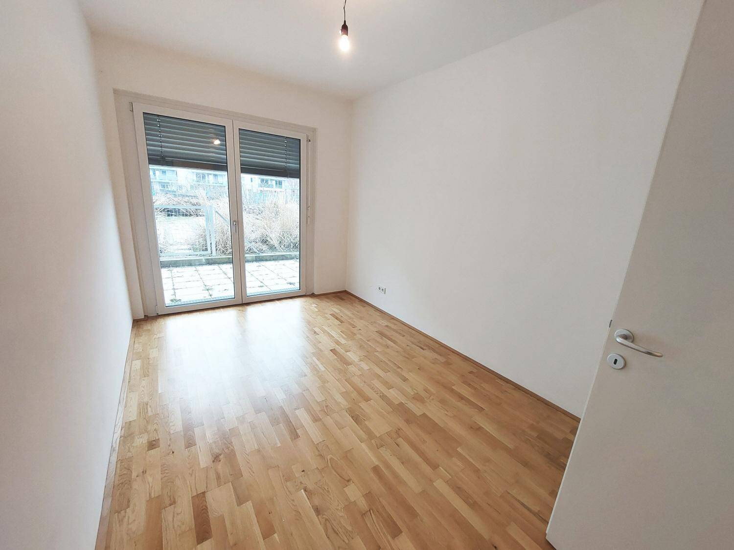 Helles Zimmer mit Parkettboden, weißen Wänden und großem Fenster mit Rollläden und Terrassenzugang.