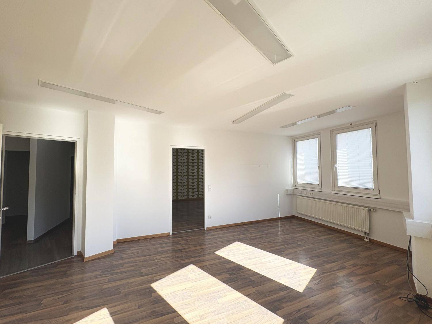 Heller, leerer Raum mit Holzboden, mehreren Fenstern und Deckenbeleuchtung.