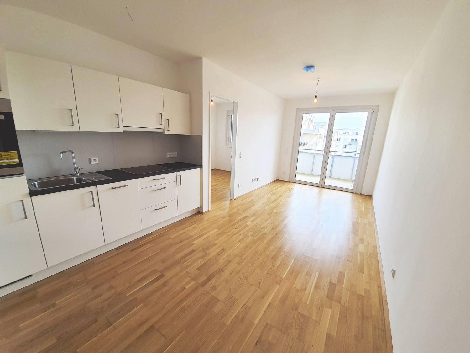 Heller offener Wohnbereich mit integrierter Küchenzeile, Parkettboden und Zugang zum Balkon.