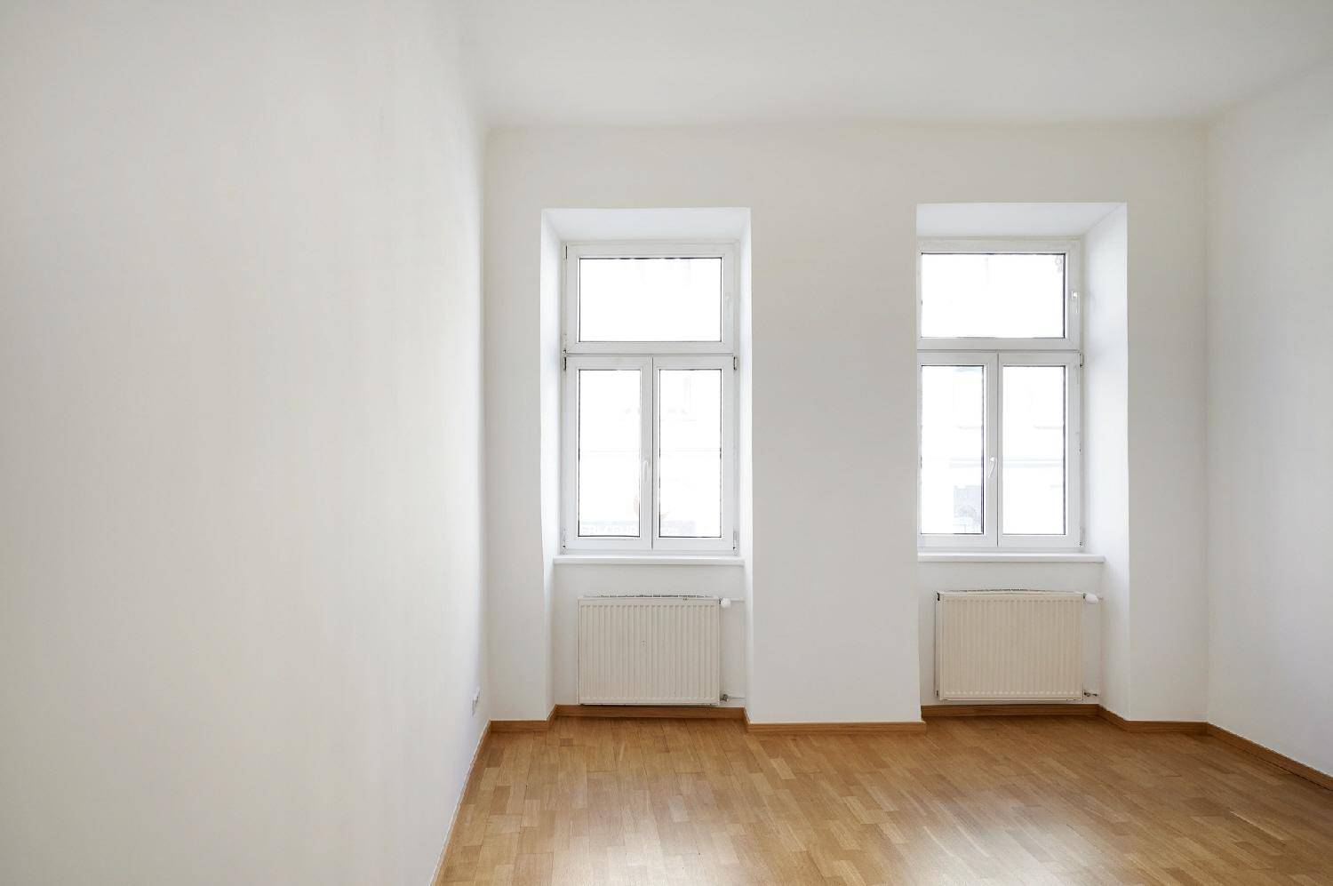 Leerer Raum mit hellen Wänden, Parkettboden und zwei großen Fenstern, ideal für Wohnzwecke.