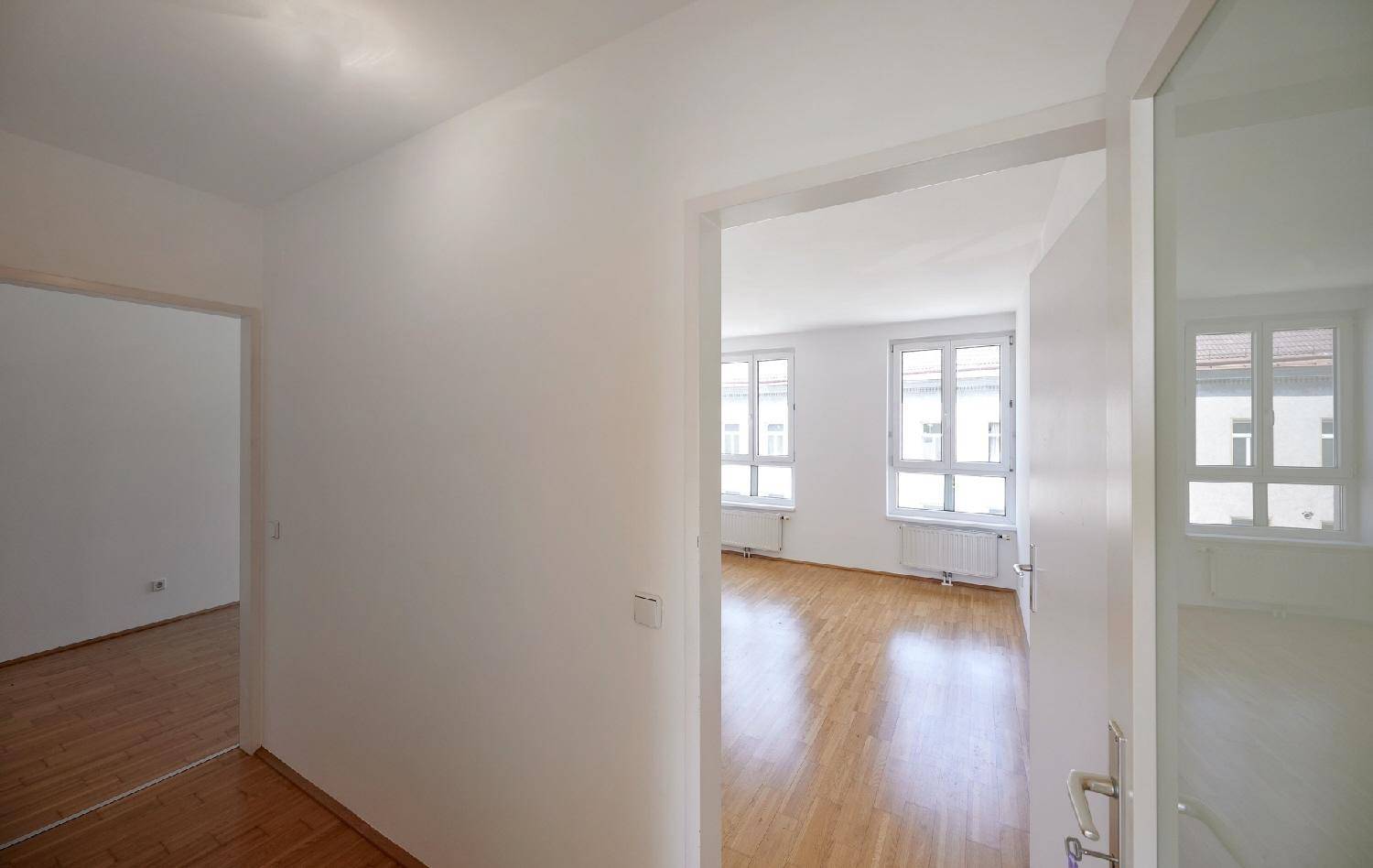 Heller Eingangsbereich mit Holzboden, der zu einem weiteren Zimmer mit Fenstern führt.