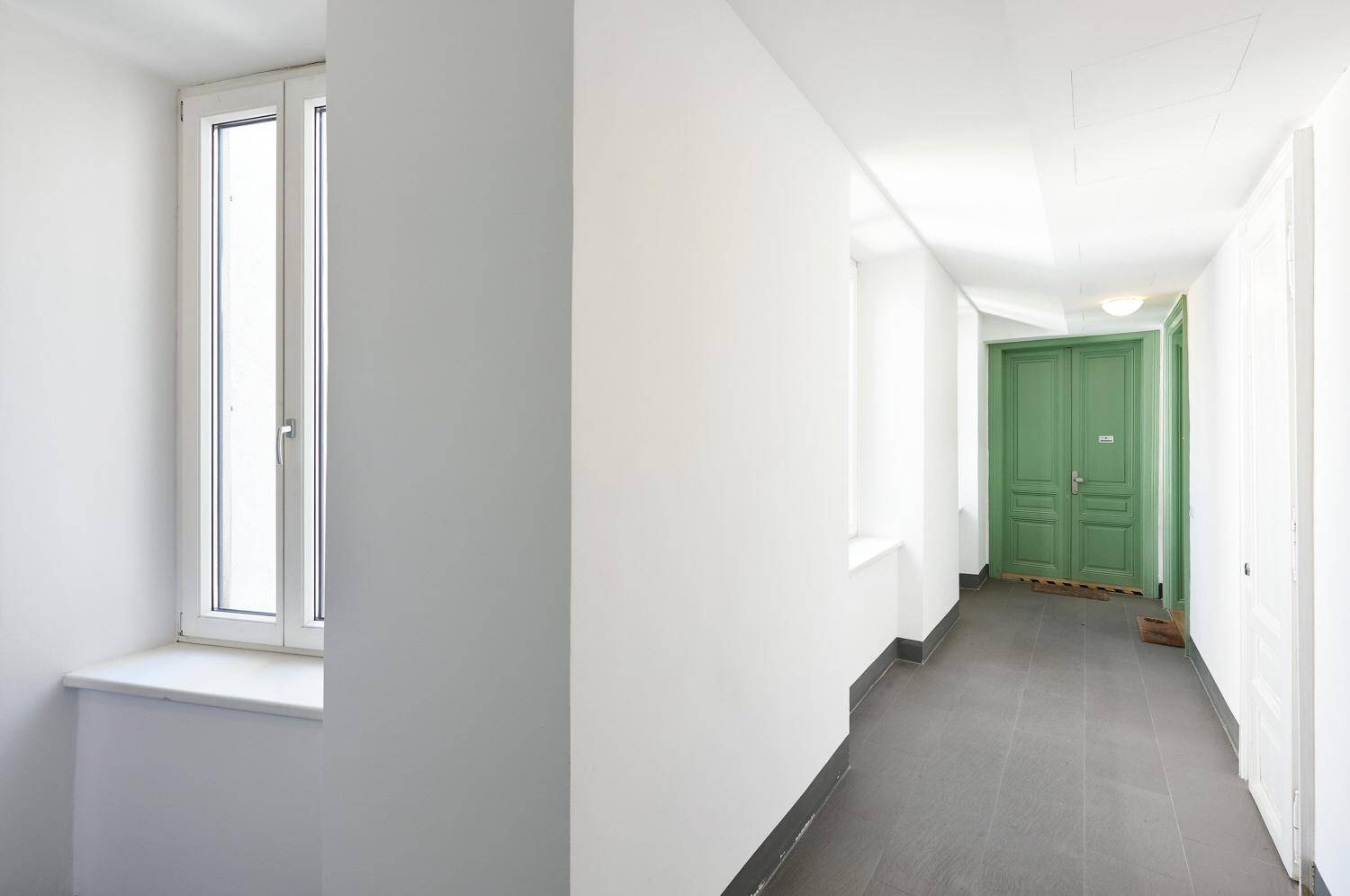 Heller Flur mit Fenster, dunklem Fliesenboden und grünen Türen.