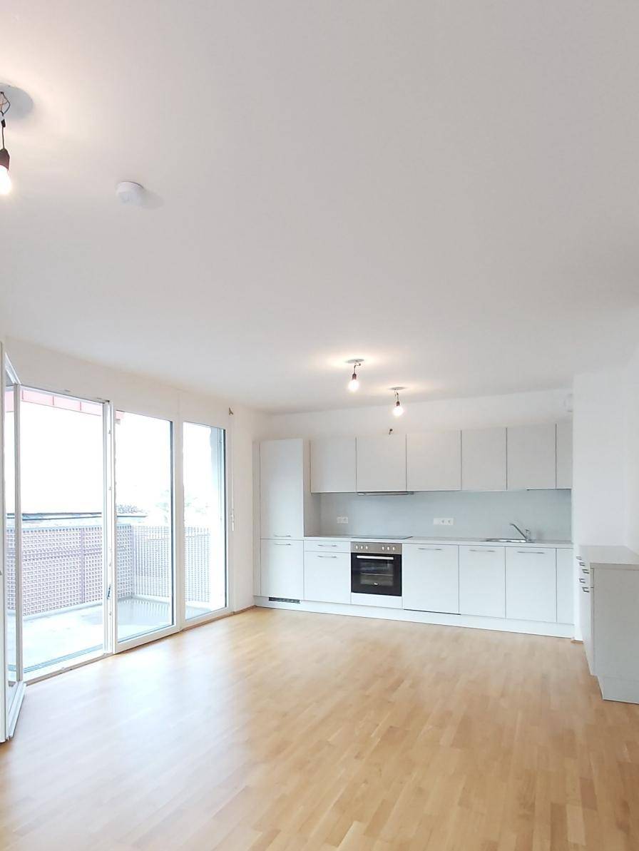 Moderner Wohnbereich mit integrierter weißer Küche, Parkettboden und viel Tageslicht durch große Fenster.