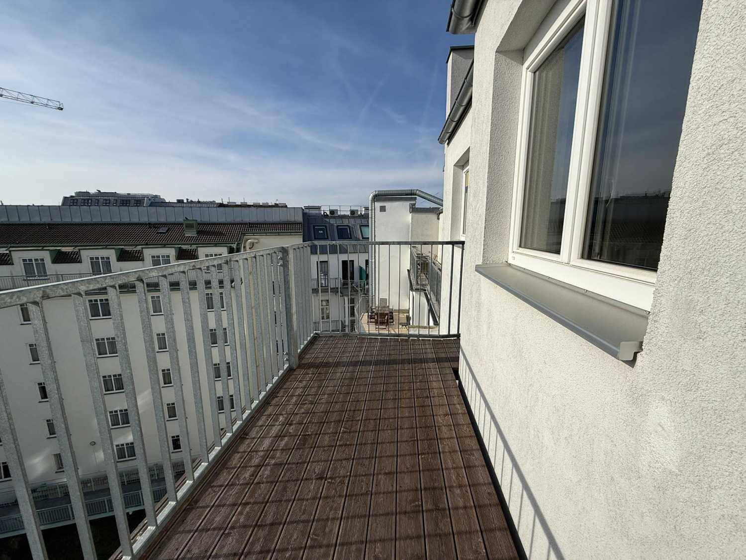 Langer Balkon mit Holzboden und Metallgeländer, bietet einen Blick auf die umliegenden Gebäude.