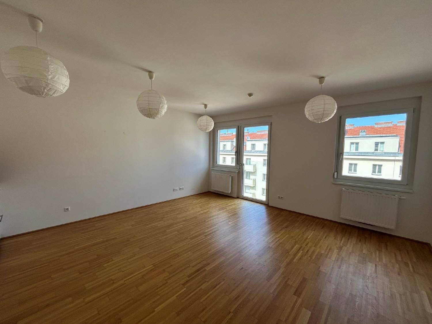 Geräumiges Wohnzimmer mit Parkettboden, großen Fenstern und Blick auf die umliegenden Gebäude.