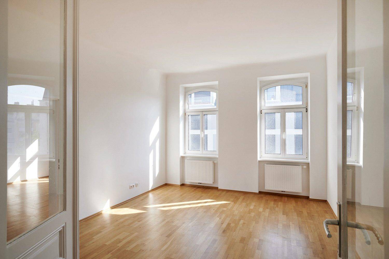 Helles Zimmer mit Parkettboden und zwei großen Fenstern, das viel Tageslicht hereinlässt.