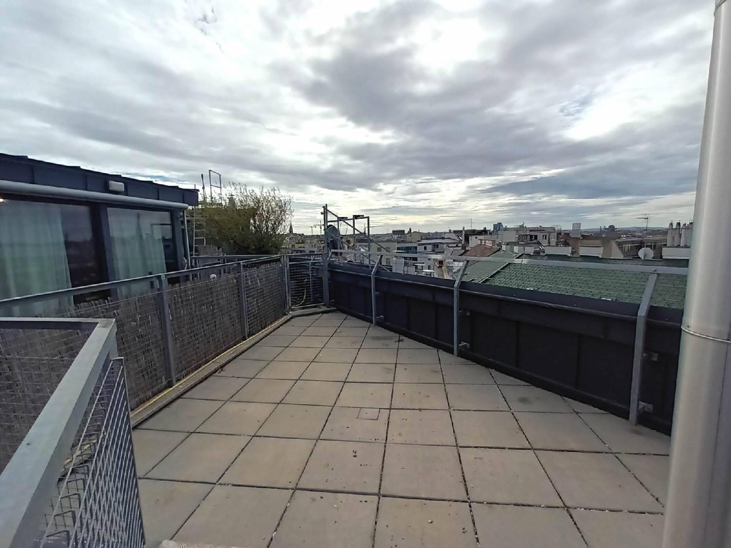 Geräumige Dachterrasse mit Fliesenboden und weitem Blick über die Dächer der Stadt unter bewölktem Himmel.