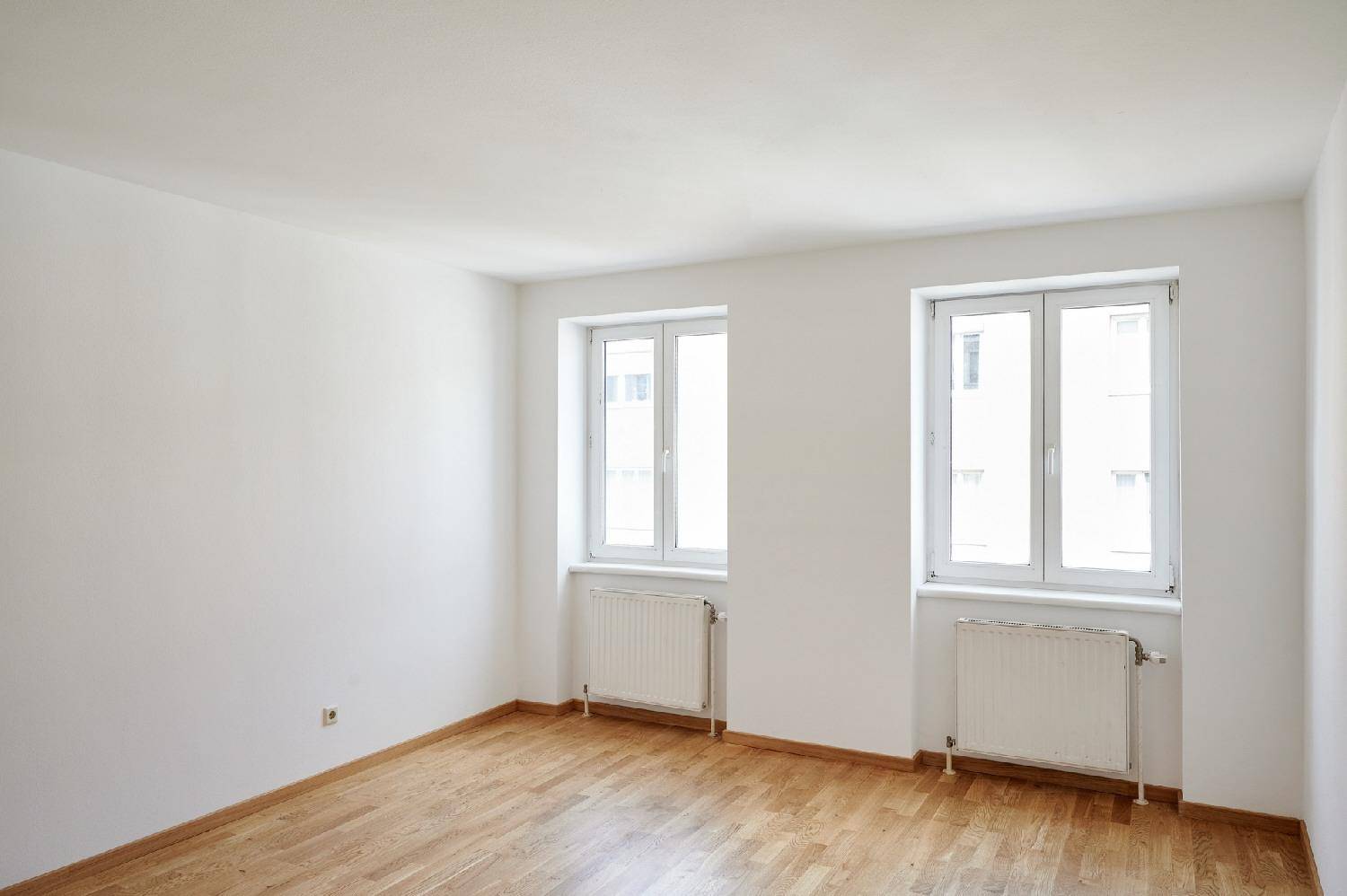 Geräumiges, helles Zimmer mit Holzboden und zwei Fenstern, ideal für Wohn- oder Schlafbereich.