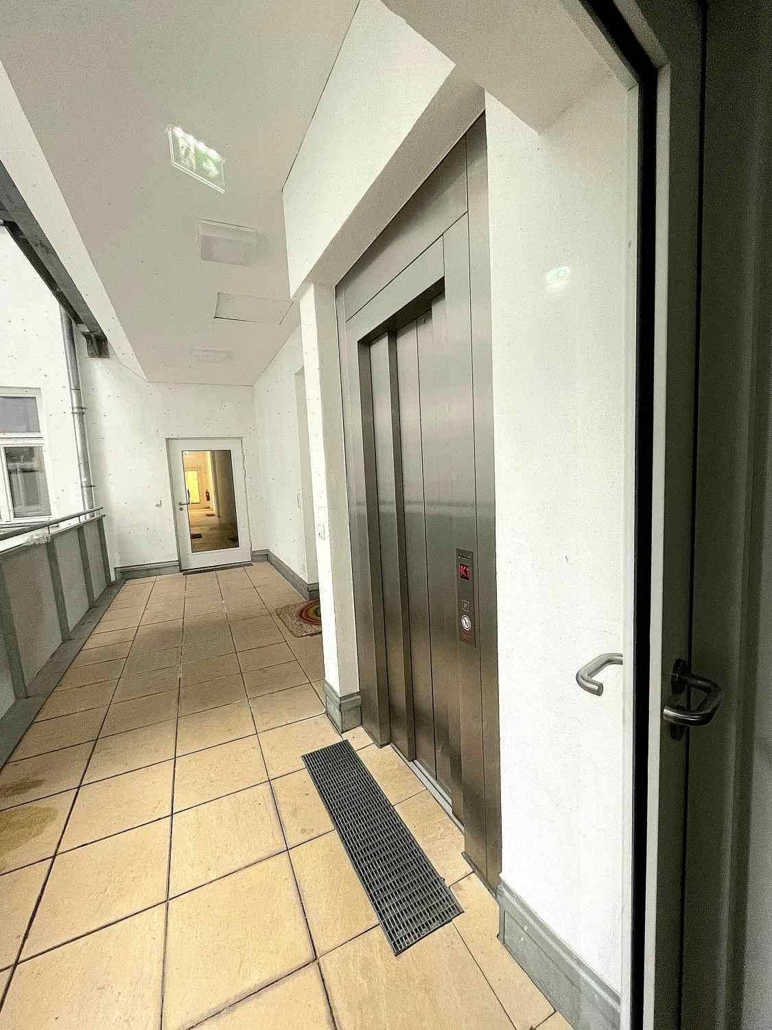 Moderner Aufzug in einem hellen Flur mit Fliesenboden, der zu den oberen Etagen führt.