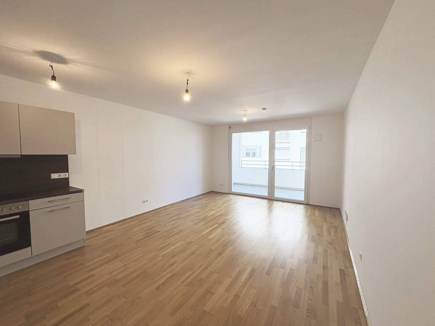 Lichtdurchfluteter Wohnbereich mit Holzboden und Zugang zu einem Balkon, inklusive Küchenzeile.