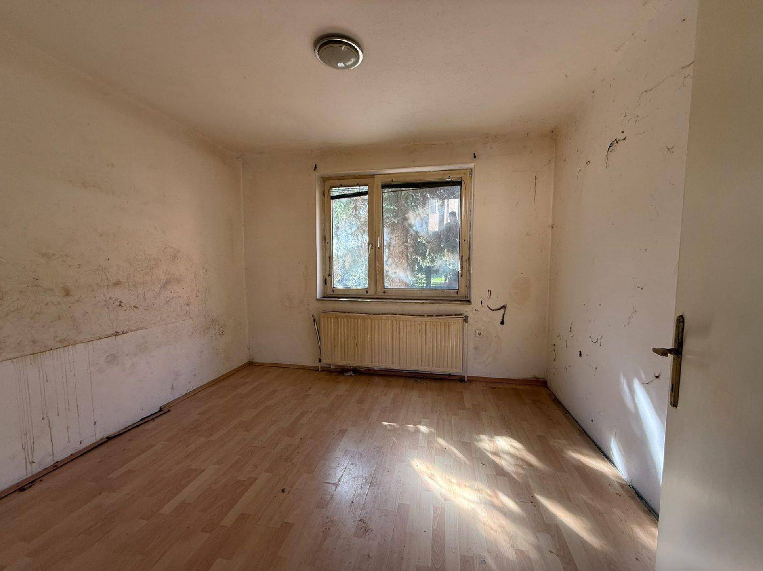 Helles Zimmer mit Fenster und Heizkörper, die Wände zeigen deutliche Gebrauchsspuren und Feuchtigkeit.