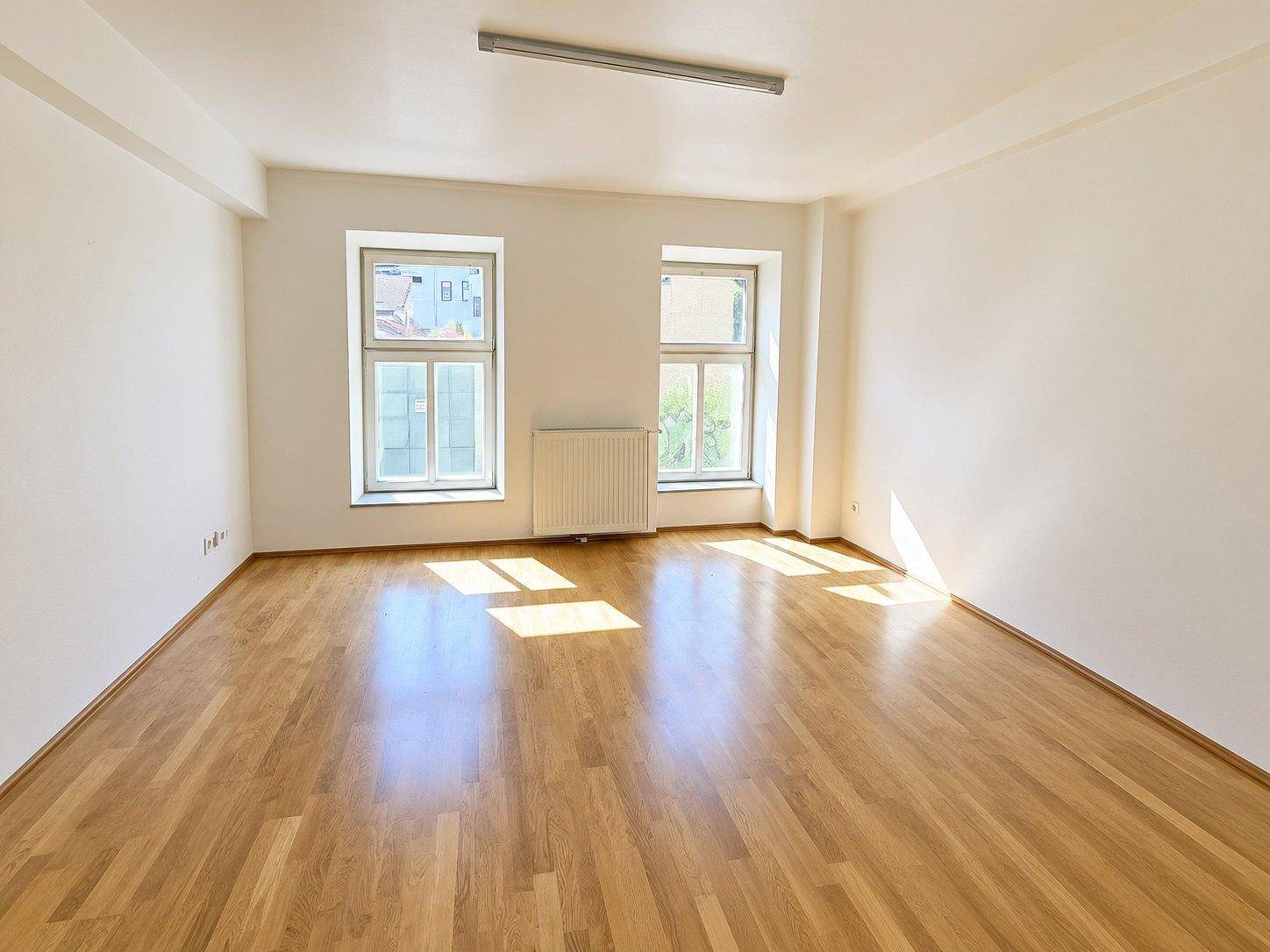 Geräumiges, helles Zimmer mit Holzboden und Heizkörper unter den Fenstern, ideal für Wohnzwecke.