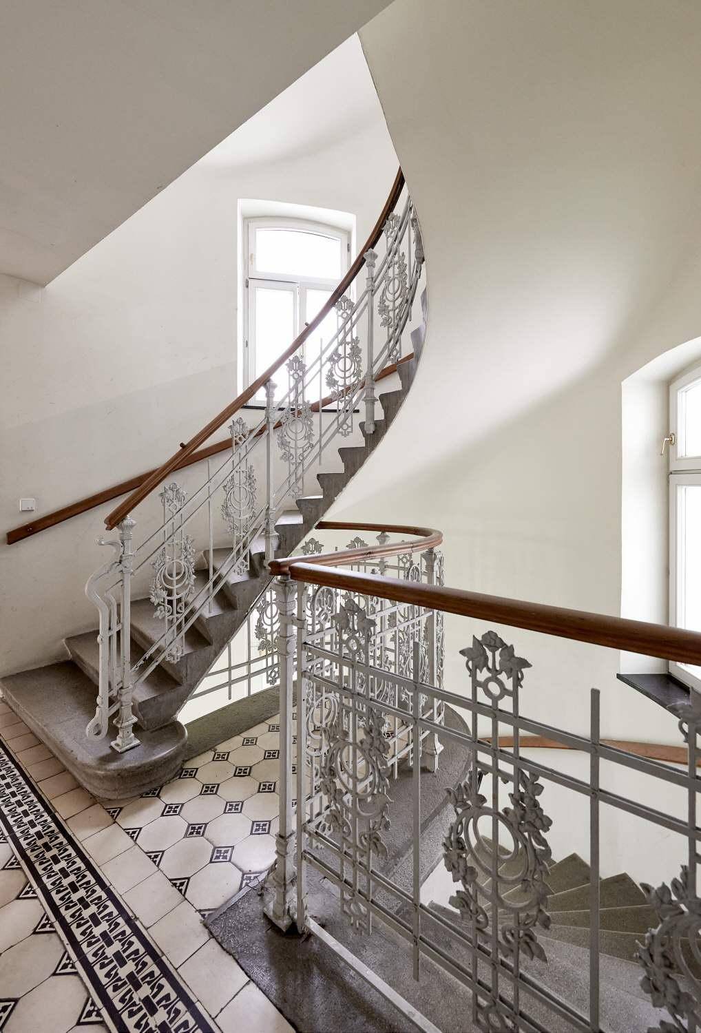 Historisches Treppenhaus mit kunstvollem Metallgeländer und gemusterten Fliesen.