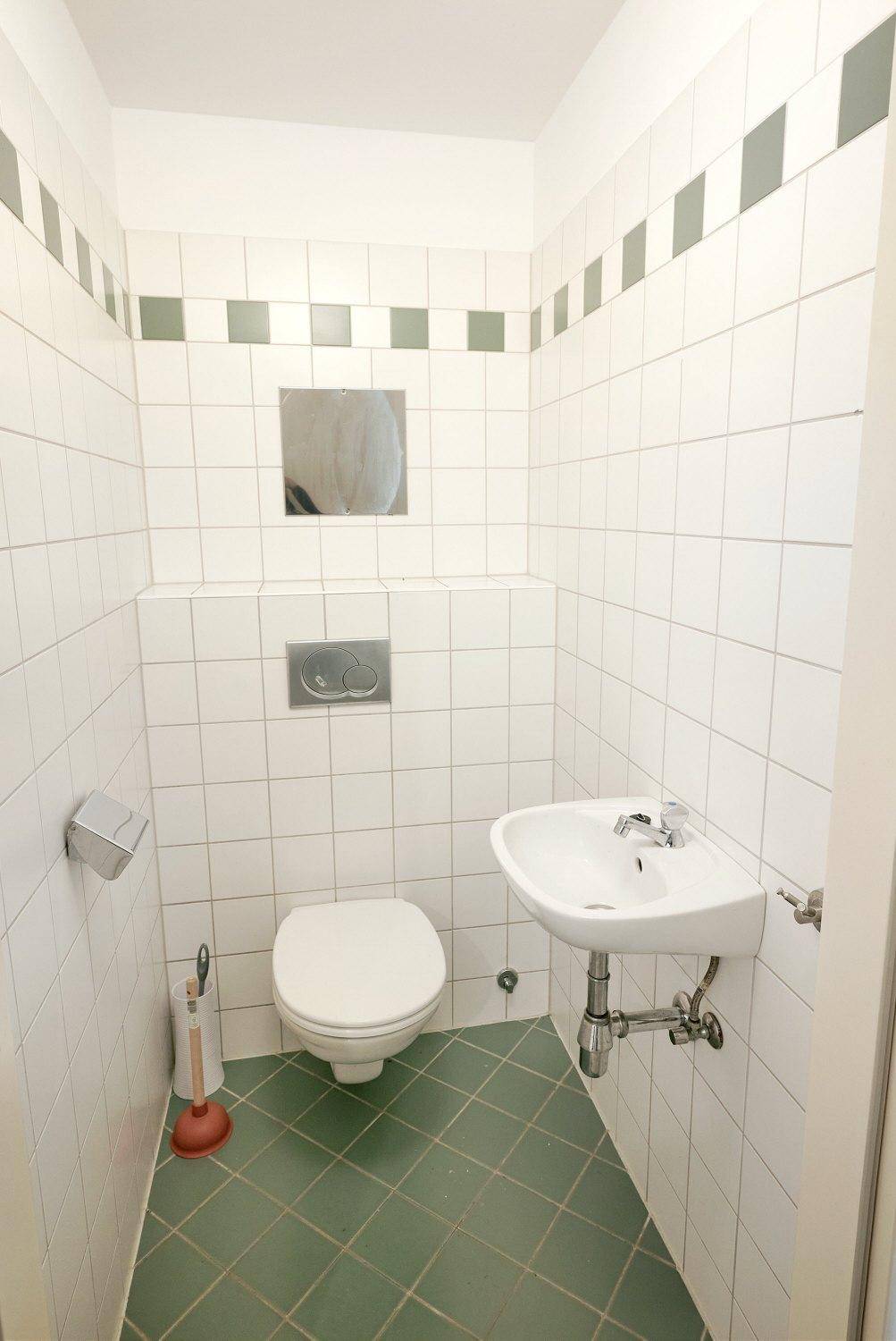 Separate Toilette mit weißen Wandfliesen, grünem Fliesenboden und einem kleinen Handwaschbecken.
