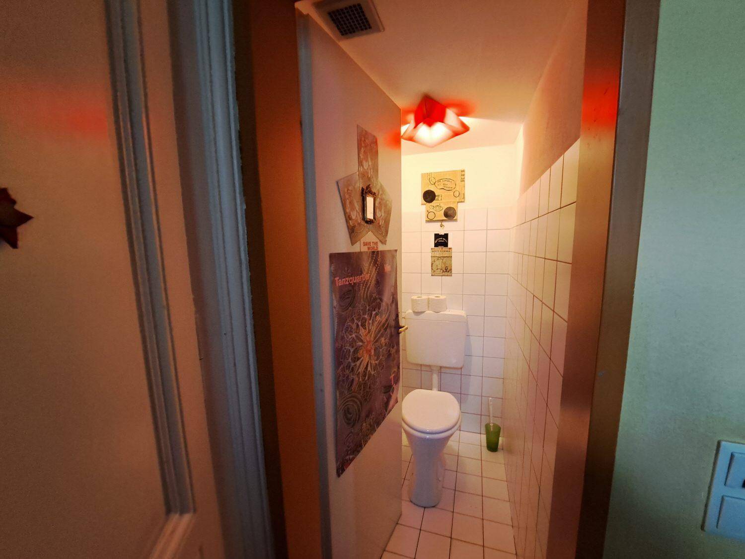 Kleines WC mit weißen Fliesen, Toilette und einer auffälligen roten Deckenleuchte, sauber und funktional.