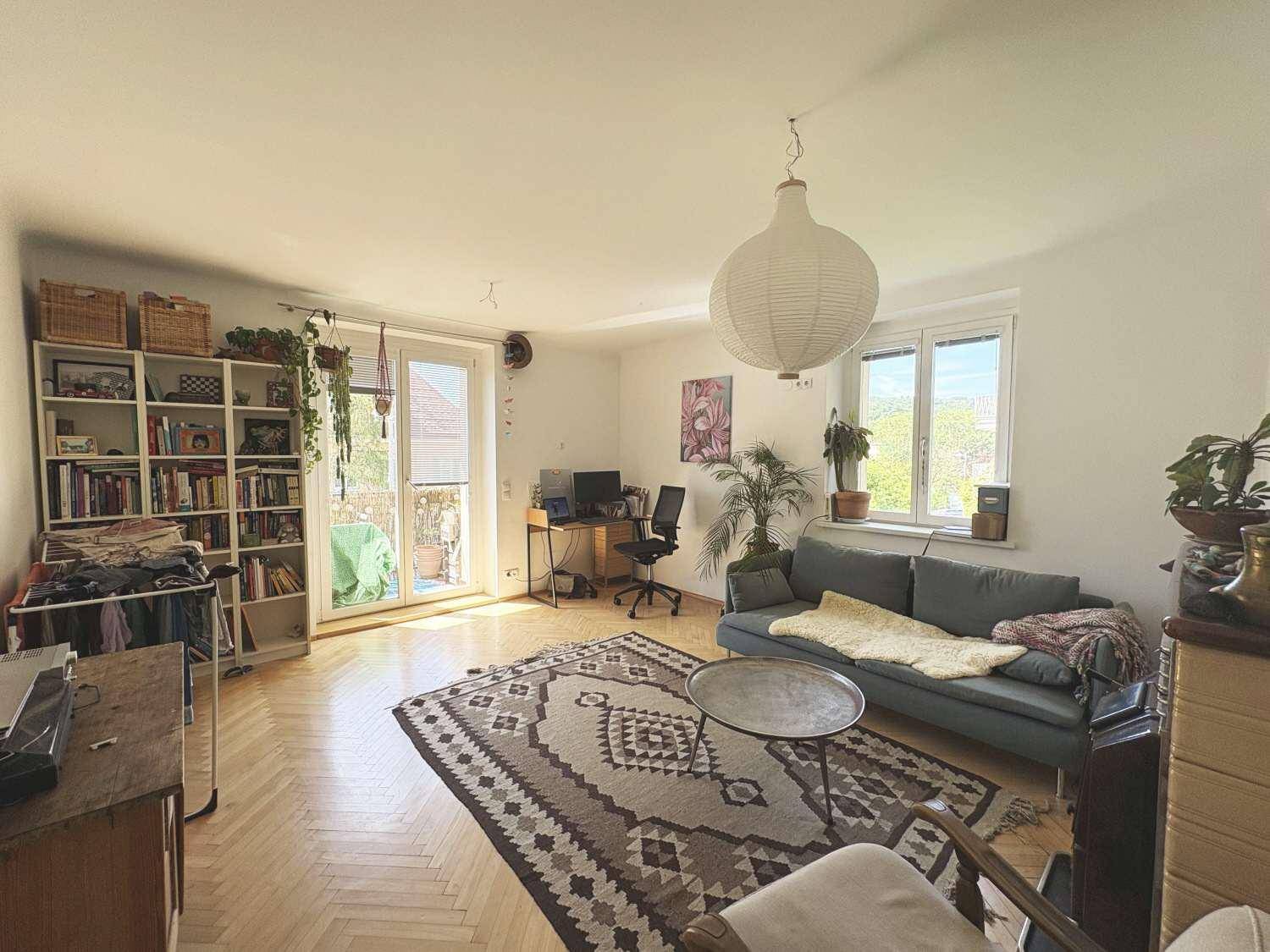 Lichtdurchflutetes Wohnzimmer mit Zugang zum Balkon, Bücherregal und Arbeitsbereich.