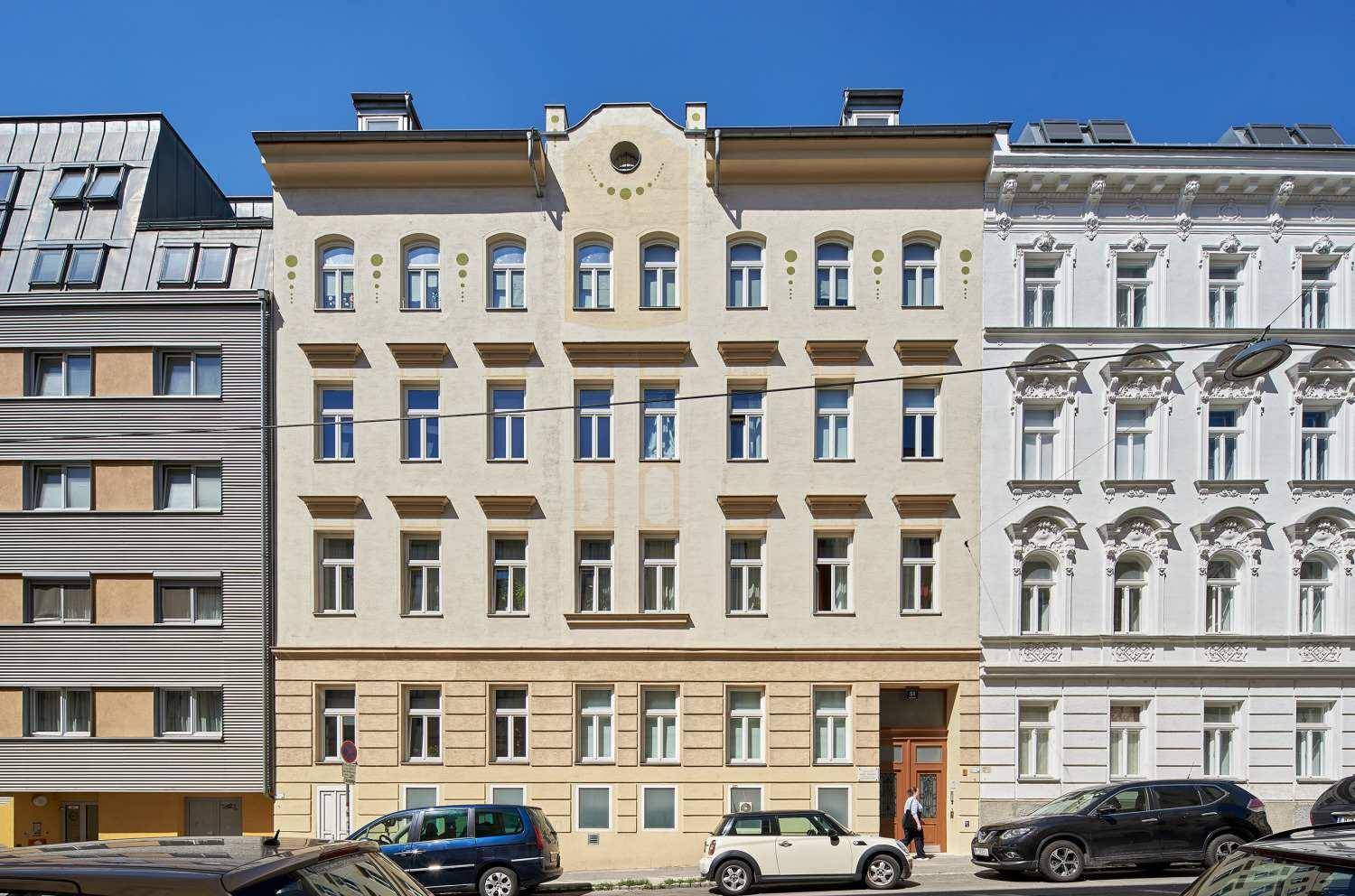 Eine symmetrische historische Gebäudefassade mit vielen Fenstern und parkenden Autos auf der Straße.